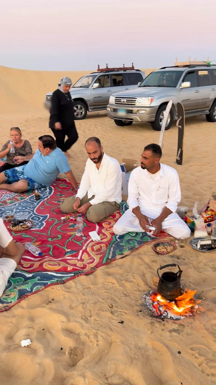 ضيوفٌ يجلسون على بساطٍ سيوي أحمر في الصحراء عند الغسق، وإبريق شاي على الجمر، وخلفهم عربتا دفع رباعي.