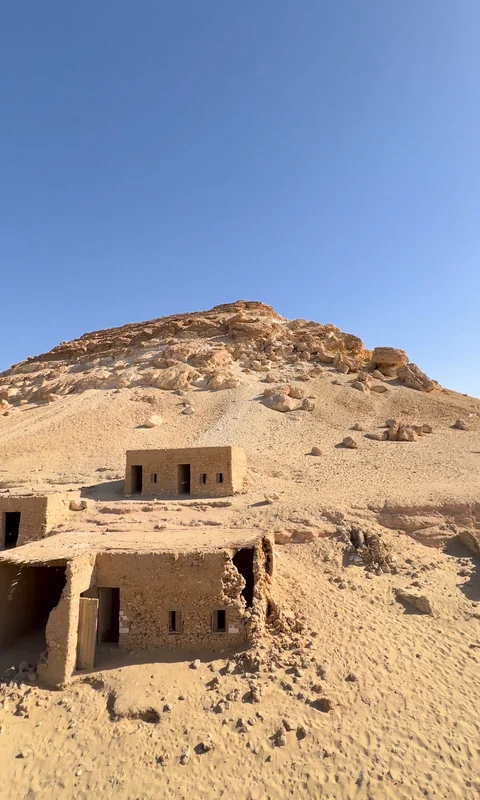 بيوتٌ صغيرة من الطين على منحدرٍ من الرمل عند جبل الدكرور، وصخور الجبل فوقها.
