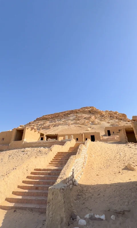 درجٌ بين جدارين من الطين يصعد إلى بيوتٍ من الطين عند سفح جبل الدكرور، والجبل خلفها تحت سماء صافية.