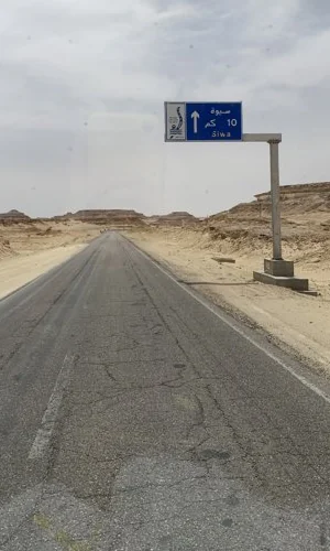 طريقٌ صحراوي خالٍ ولافتةٌ زرقاء عليه: سيوة 10 كم.