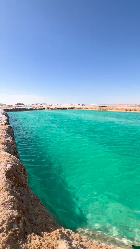 بركة ملحٍ فيروزية محفورة بين ضفافٍ من الملح والطين، تحت سماءٍ زرقاء صافية، شرق سيوة.